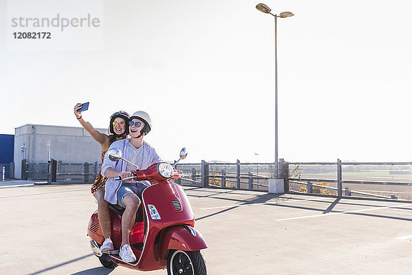 Glückliches junges Paar mit einem Selfie auf dem Motorroller auf der Parkebene