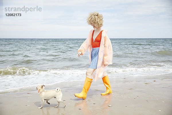 Lächelnde junge Frau in Regenmantel und Wellington-Stiefeln  die mit ihrem Hund am Meer spazieren geht.