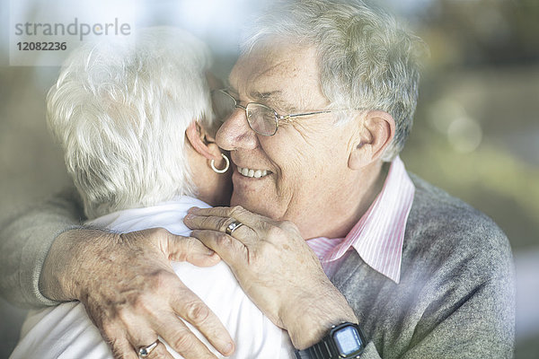 Fröhliches Seniorenpaar umarmt sich hinter der Fensterscheibe