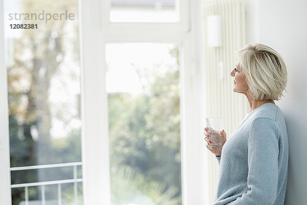 Lächelnde Seniorin mit einem Glas Wasser aus dem Fenster schauend