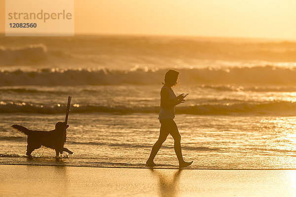 Indonesien  Bali  Silhouette einer Frau  die bei Sonnenuntergang mit ihrem Hund am Strand spazieren geht.