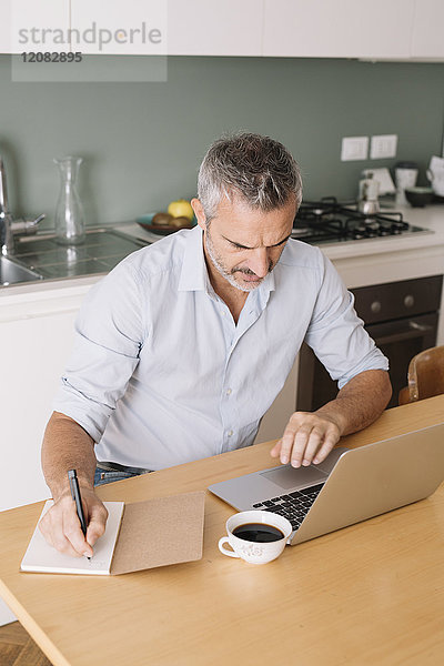 Mann schreibt in Notizbuch und benutzt Laptop im Home-Office