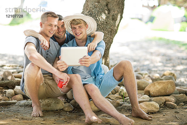 Drei junge männliche Freunde sitzen auf Felsen und schauen auf ein digitales Tablett