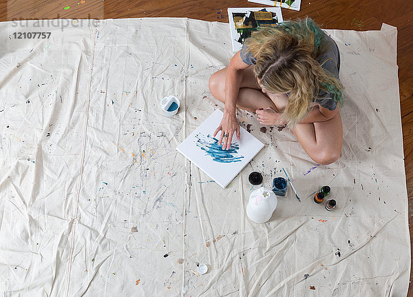 Draufsicht einer Künstlerin  die auf einem Staubtuch sitzt und Fingerfarben auf abstrakte Leinwand malt
