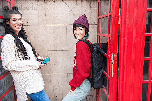 Porträt von zwei jungen stilvollen Frauen  die sich an rote Telefonzellen lehnen  London  UK