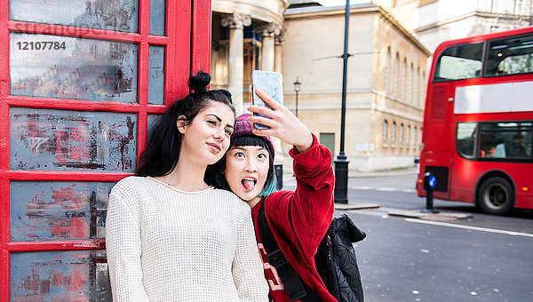 Zwei junge stilvolle Frauen beim Smartphone-Selfie an der roten Telefonzelle  London  UK