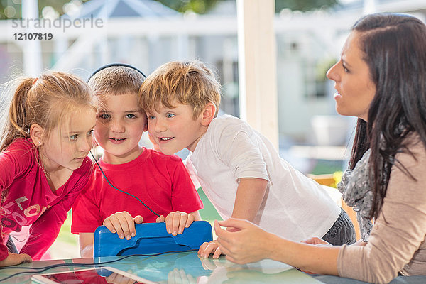Mittlere erwachsene Frau sitzt mit drei kleinen Kindern  ein Junge hält ein digitales Tablet und trägt Kopfhörer