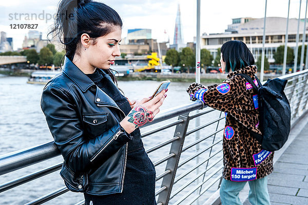 Junge Frau betrachtet Smartphone mit Freundin  die von der Millennium-Fußgängerbrücke hinaussieht  London  Großbritannien