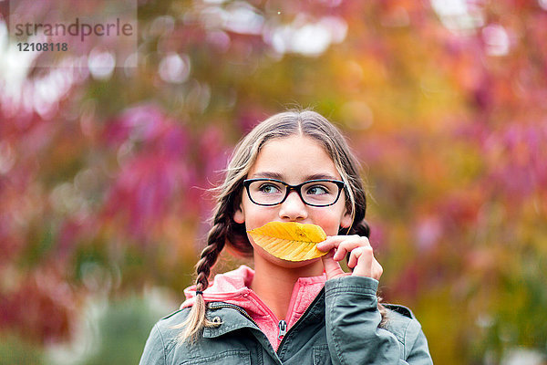 Porträt eines Mädchens mit Zöpfen und Brille  das den Mund mit einem Blatt bedeckt und weg schaut