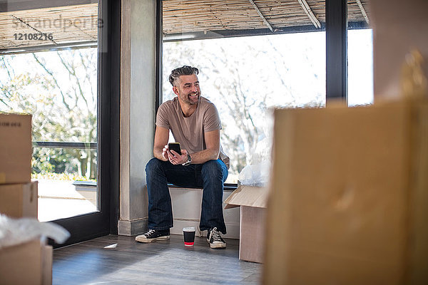 Mann sitzt in unmöbliertem Haus  umgeben von Pappkartons  und benutzt ein Smartphone