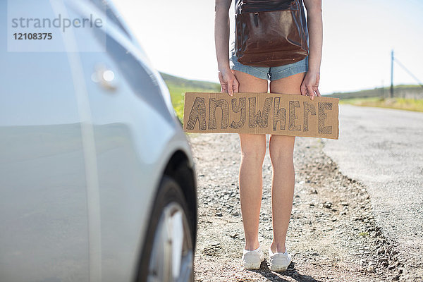 Junge Frau steht neben dem Auto und hält ein Trampschild mit der Aufschrift überall in der Hand  niedriger Abschnitt