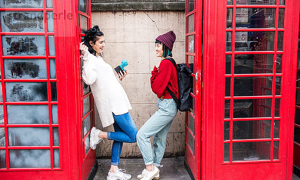 Zwei junge stilvolle Frauen  die sich an rote Telefonzellen lehnen  London  UK