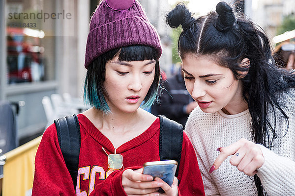 Zwei junge  stilvolle Frauen schauen sich auf der Straße in der Stadt ein Smartphone an