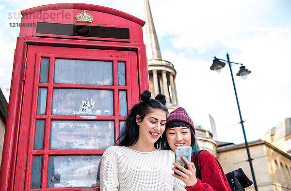 Zwei junge  stilvolle Frauen betrachten ein Smartphone an einer roten Telefonzelle  London  UK