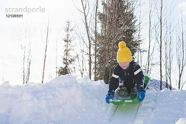 Junge mit gelber Strickmütze beim Schlittenfahren auf schneebedecktem Hügel