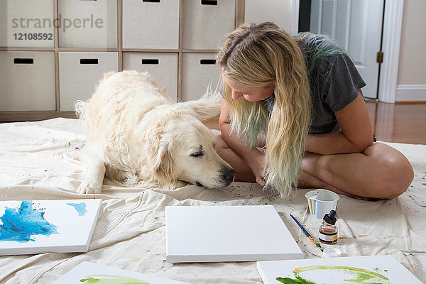 Künstlerin sitzt auf einem Staubtuch und streichelt Hund im Atelier