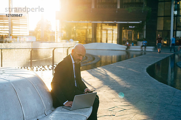 Reifer Geschäftsmann sitzt im Freien  benutzt Laptop  hält Smartphone