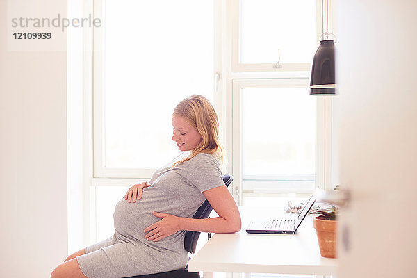 Schwangere junge Frau am Schreibtisch mit Blick auf den Bauch