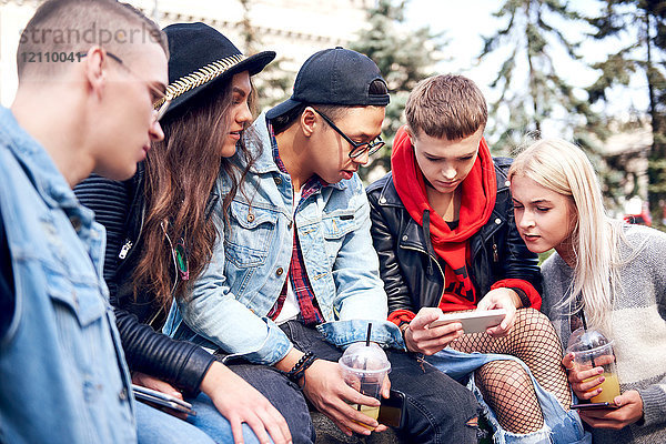 Fünf junge erwachsene Freunde sitzen in der Stadt an der Wand und schauen auf ein Smartphone