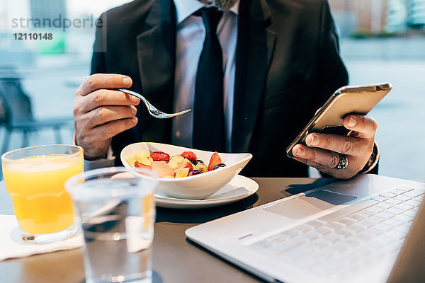 Reifer Geschäftsmann sitzt im Freien  frühstückt  benutzt Smartphone  Laptop auf dem Tisch  Mittelteil