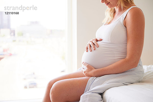 Schnappschuss einer schwangeren jungen Frau  die mit den Händen auf dem Bauch auf dem Bett sitzt