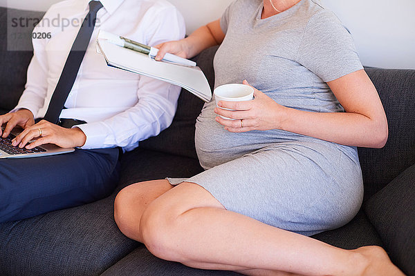 Nackenansicht eines schwangeren Paares  das mit Zeitschrift und Laptop auf dem Sofa sitzt