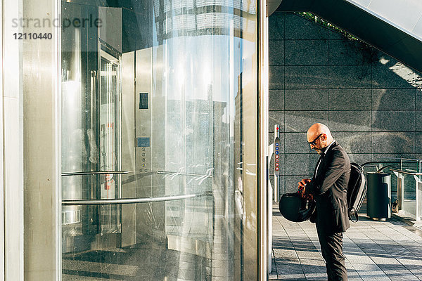 Reifer Geschäftsmann im Freien  wartet auf den Aufzug  kontrolliert die Armbanduhr  hält den Motorradhelm