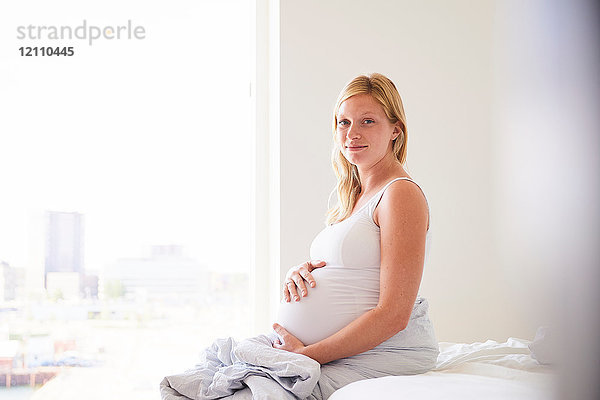 Porträt einer schwangeren jungen Frau  die mit den Händen auf dem Bauch auf dem Bett sitzt