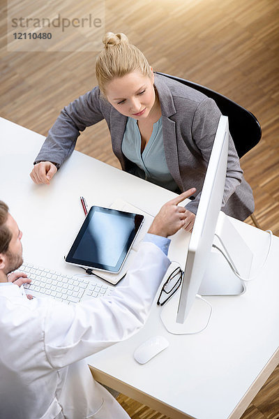 Männlicher Arzt und junge Frau sitzen am Tisch und schauen auf den Computerbildschirm