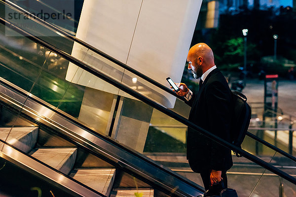 Reifer Geschäftsmann nachts im Freien  auf der Rolltreppe  mit Smartphone