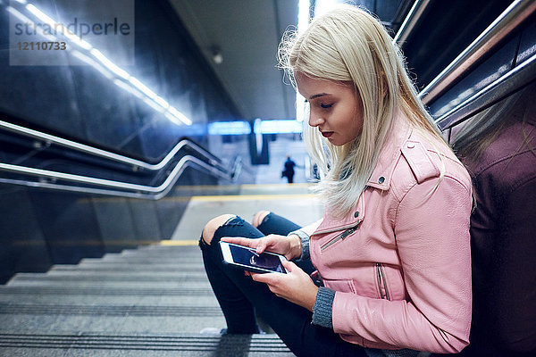 Junge Frau sitzt auf der Treppe der U-Bahn-Station und schaut auf ein Smartphone