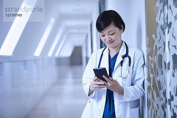 Arzt im Krankenhausflur lehnt mit Smartphone an Wand