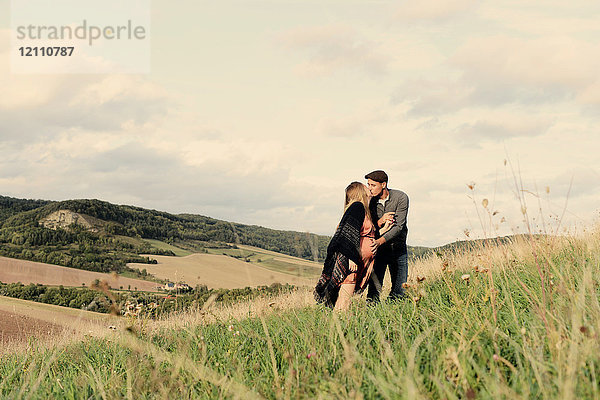 Romantisches schwangeres Paar mittlerer Erwachsener küsst sich am ländlichen Hang