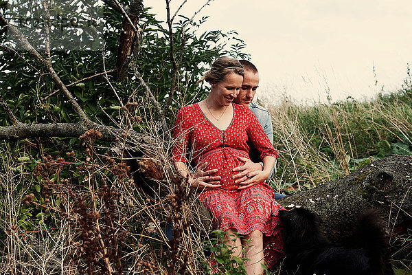 Romantisches schwangeres Paar im mittleren Erwachsenenalter auf einem Baumstamm sitzend