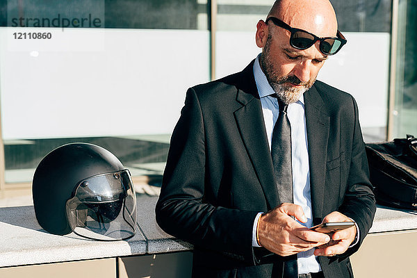 Älterer Geschäftsmann steht im Freien  benutzt Smartphone  Motorradhelm an der Wand neben ihm