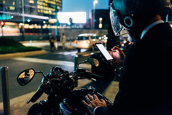 Älterer Geschäftsmann nachts im Freien  auf dem Motorrad sitzend  Motorradhelm tragend  Smartphone benutzen  Rückansicht