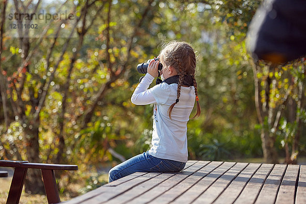 Mädchen in ländlicher Umgebung  sitzt auf einer Terrasse und schaut durch ein Fernglas