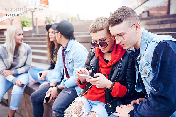 Fünf junge erwachsene Freunde schauen auf Smartphones und unterhalten sich auf der Stadttreppe