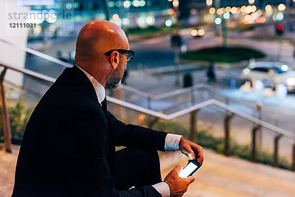 Ein reifer Geschäftsmann  der nachts im Freien auf einer Treppe sitzt und sein Smartphone in der Hand hält
