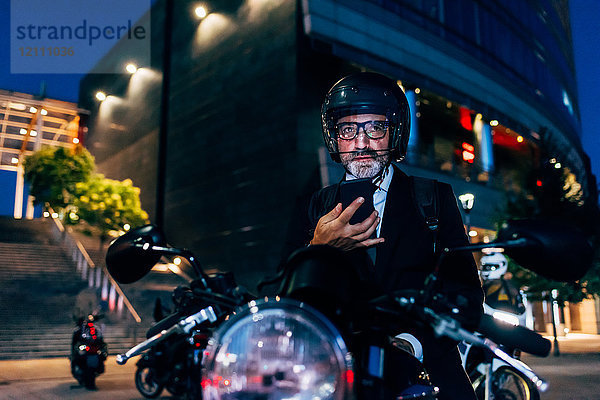 Reifer Geschäftsmann nachts im Freien  sitzt auf einem Motorrad und benutzt ein Smartphone