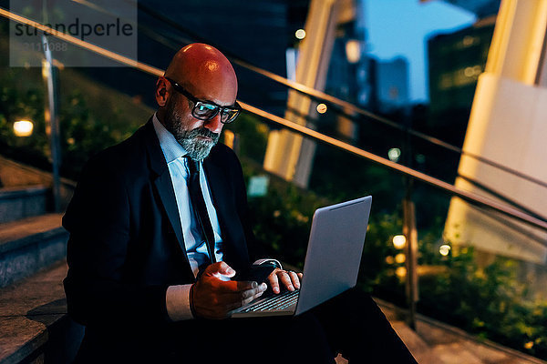 Reifer Geschäftsmann nachts im Freien  sitzt auf Treppen  benutzt Laptop  hält Smartphone