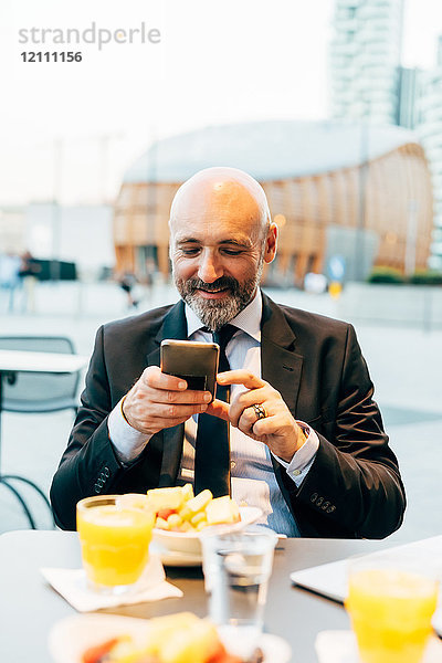 Reifer Geschäftsmann sitzt draußen im Café und benutzt ein Smartphone