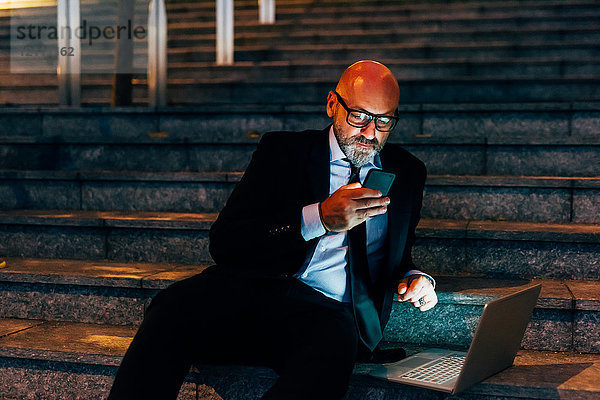 Älterer Geschäftsmann nachts im Freien  auf der Treppe sitzend  mit Smartphone  Laptop auf der Stufe neben ihm
