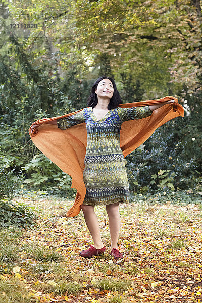 Volle Länge der Frau mit orangem Schal stehend mit ausgestreckten Armen im Park im Herbst