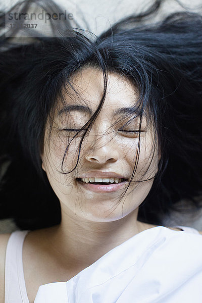 Hochwinkelansicht einer lächelnden jungen Frau mit geschlossenen Augen und unordentlichem Haar auf dem Bett liegend.
