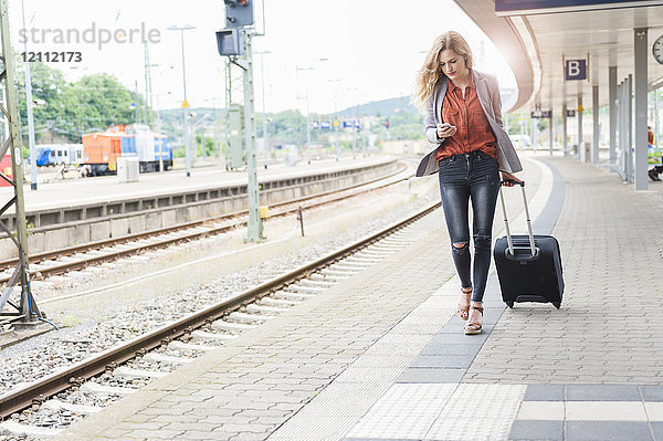 Junge Frau mit Rollgepäck schaut auf ihr Smartphone  während sie auf dem Bahnsteig spazieren geht.