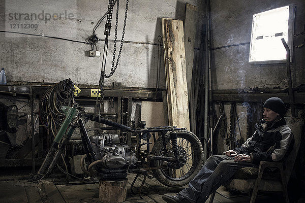 Mechaniker sitzt im Sessel und raucht Zigarette in der Werkstatt mit zerlegtem Oldtimer-Motorrad