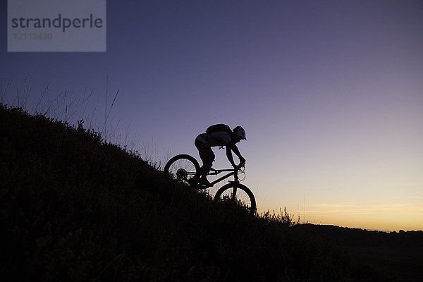 Männlicher Mountainbiker mit Silhouette  der bei Sonnenuntergang den Berg hinunterfährt