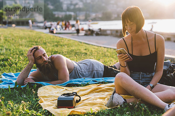 Junges Paar betrachtet Smartphone im Gras am Wasser  Comer See  Lombardei  Italien