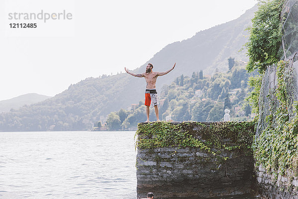 Junger männlicher Schwimmer steht mit offenen Armen am Pier  Comer See  Lombardei  Italien